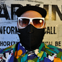 Man wearing black mask with white sunglasses and colorful shirt. Background has painted letters.