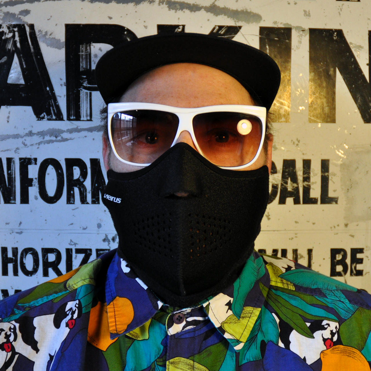 Man wearing black mask with white sunglasses and colorful shirt. Background has painted letters.