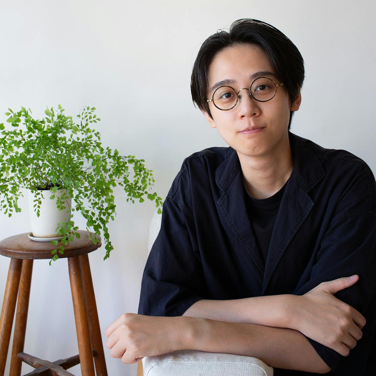 A person with short black hair sitting with their arms crossed next to a plant.